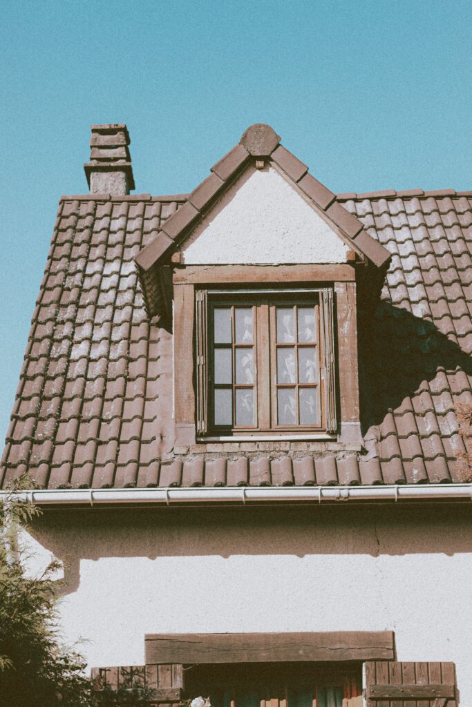 Low angle of tiled roof of stylish rustic cottage with attic window and chimney against cloudless blue sky